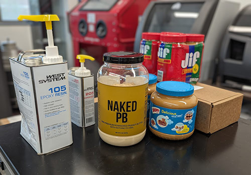 Several containers of epoxy alongside jars of peanut butter and peanut butter powder sitting on a table.